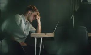 Ein Mann sitzt mit der Hand an der Stirn vor einem Laptop. Er wirkt nachdenklich, aber es ist möglich, dass er trotz Depressionen arbeitet.