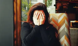 Frau mit roten Haaren verdeckt ihr Gesicht mit den Händen, als würde sie grundlos weinen. Sie sitzt in einem Sessel mit geometrischem Muster.