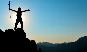 Silhouette einer Person mit Wanderstöcken auf einem Berggipfel, die ihre Arme ausbreitet. Ein Symbol für das Erreichen von Zielen im Leben.