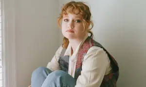 Nachdenkliche junge Frau mit roten Haaren, die sich fragt, was sind minderwertigkeitskomplexe. Sie sitzt in Jeans und einer Weste vor einer hellen Wand.