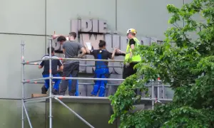 Arbeiter auf Gerüst montieren Buchstaben an einer Fassade. Ein Mensch in Warnweste gibt Anweisungen.