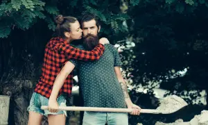 Ein Mann mit Bart und eine Frau mit Zopf, die ihm einen Kuss auf die Wange gibt, halten eine Schaufel. Sie genießen ihren **Freiraum in einer Beziehung** im Grünen.