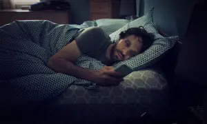 Mann liegt im Bett, starrt auf sein Handy und scrollt durch negative Nachrichten. Ein klassischer Fall von Doomscrolling.