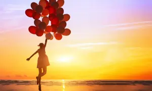 Mädchen mit vielen roten und orangen Ballons springt am Strand in den Sonnenuntergang. Sie genießt es, ganz sie selbst zu sein.