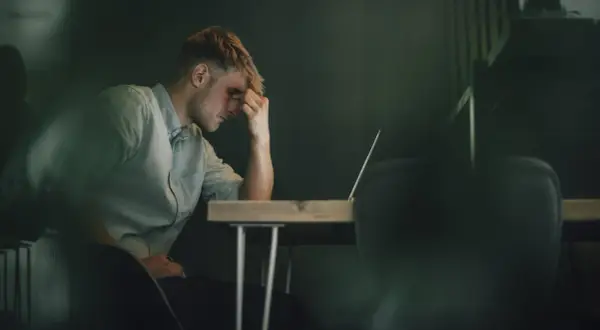 Ein Mann sitzt mit der Hand an der Stirn vor einem Laptop. Er wirkt nachdenklich, aber es ist möglich, dass er trotz Depressionen arbeitet.