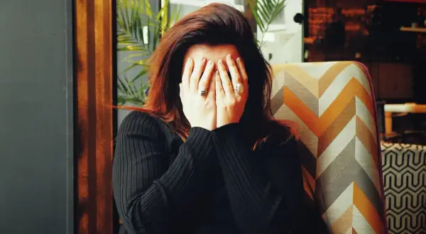 Frau mit roten Haaren verdeckt ihr Gesicht mit den Händen, als würde sie grundlos weinen. Sie sitzt in einem Sessel mit geometrischem Muster.
