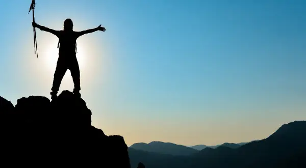 Silhouette einer Person mit Wanderstöcken auf einem Berggipfel, die ihre Arme ausbreitet. Ein Symbol für das Erreichen von Zielen im Leben.
