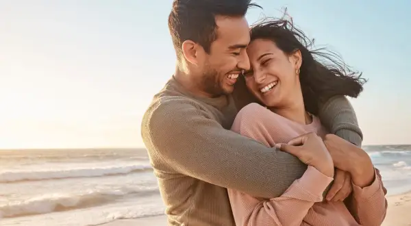 Glückliches Paar umarmt sich am Strand, lacht und genießt die Zeit. Ein perfekter Moment, um die Beziehung aufzufrischen.