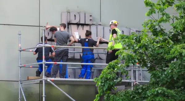 Arbeiter auf Gerüst montieren Buchstaben an einer Fassade. Ein Mensch in Warnweste gibt Anweisungen.