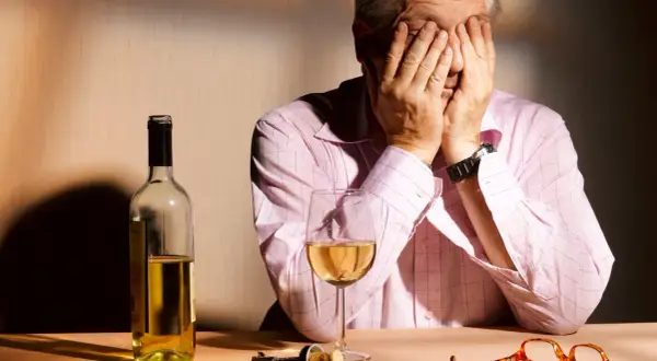 Mann mit Händen vor dem Gesicht, Wein, Korkenzieher und Brille auf dem Tisch. Ein Bild, das die Verzweiflung und die Folgen von Korsakow-Syndrom symbolisiert.