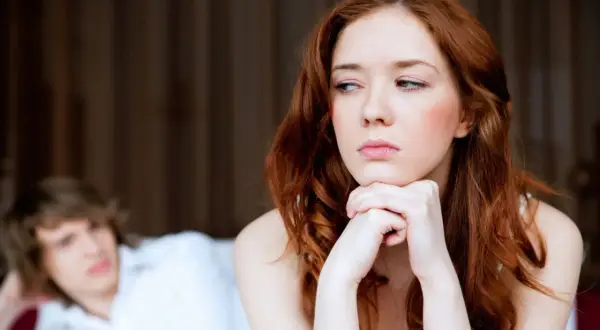 Traurige Frau mit roten Haaren blickt weg. Hinter ihr liegt ein Mann im Bett. Sie spürt keine sexuelle Anziehung mehr zum Partner.