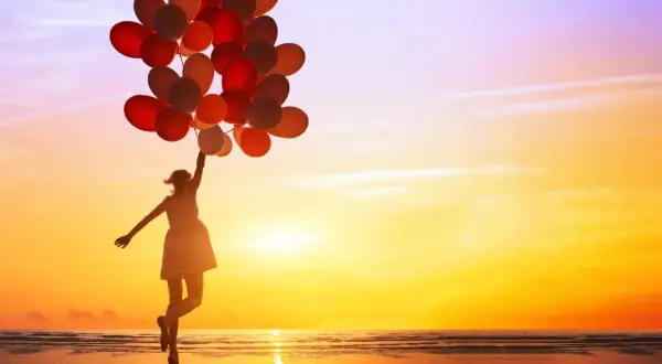Mädchen mit vielen roten und orangen Ballons springt am Strand in den Sonnenuntergang. Sie genießt es, ganz sie selbst zu sein.
