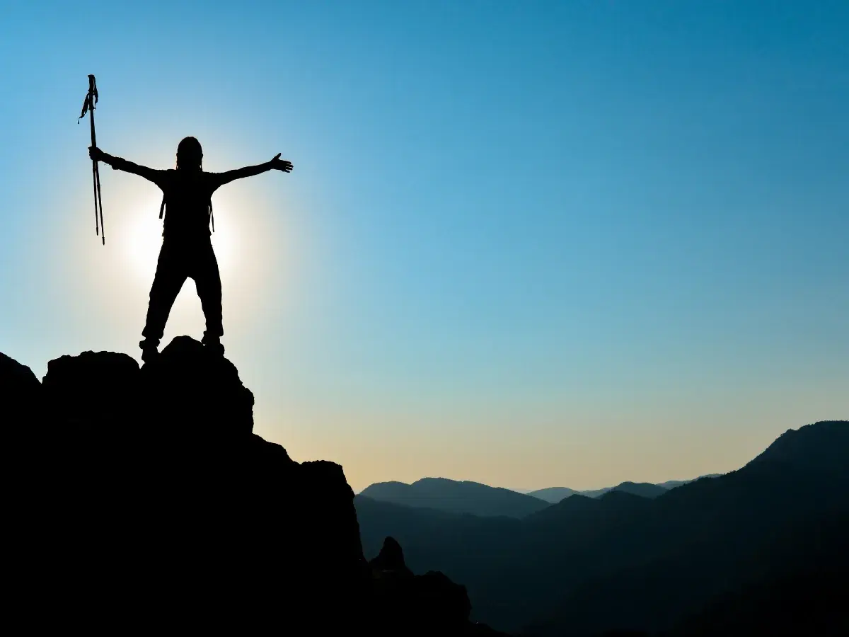 Silhouette einer Person mit Wanderstöcken auf einem Berggipfel, die ihre Arme ausbreitet. Ein Symbol für das Erreichen von Zielen im Leben.