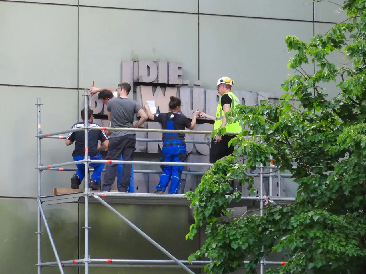 Arbeiter auf Gerüst montieren Buchstaben an einer Fassade. Ein Mensch in Warnweste gibt Anweisungen.