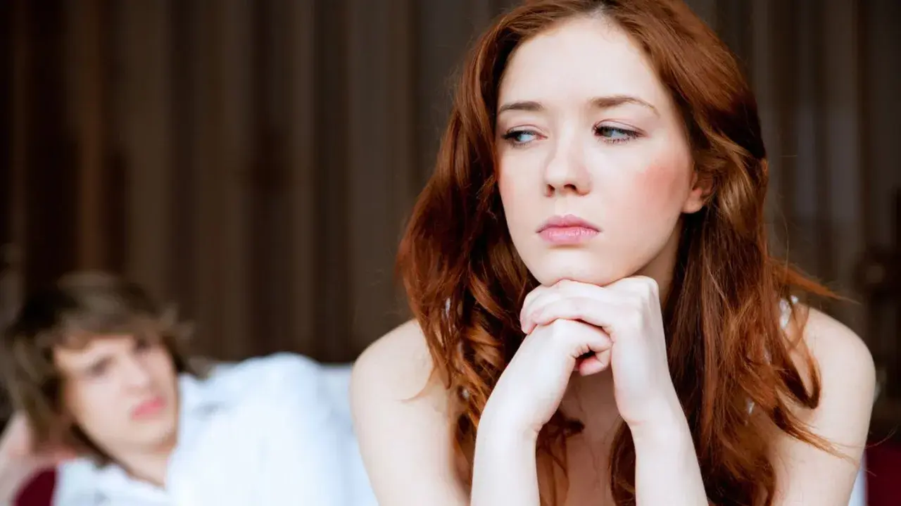 Traurige Frau mit roten Haaren blickt weg. Hinter ihr liegt ein Mann im Bett. Sie spürt keine sexuelle Anziehung mehr zum Partner.