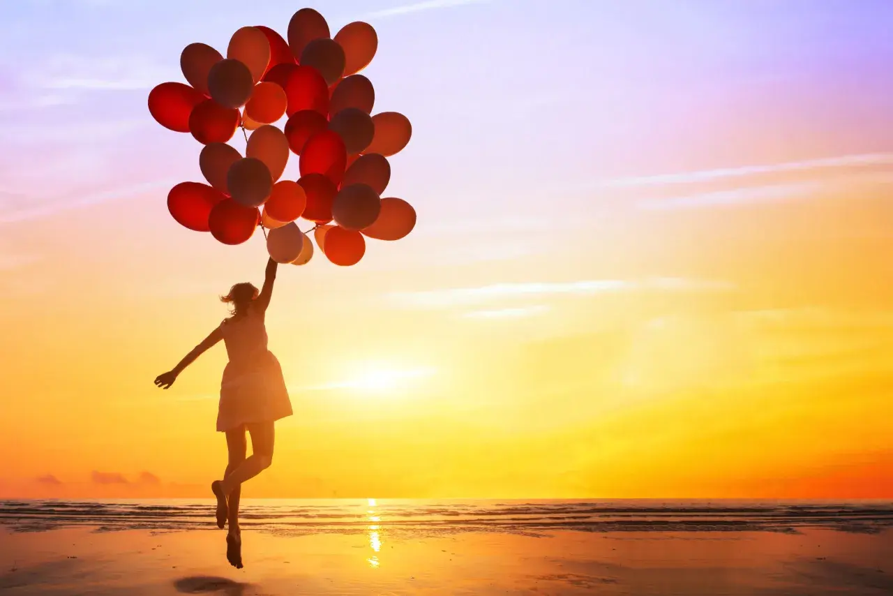 Mädchen mit vielen roten und orangen Ballons springt am Strand in den Sonnenuntergang. Sie genießt es, ganz sie selbst zu sein.