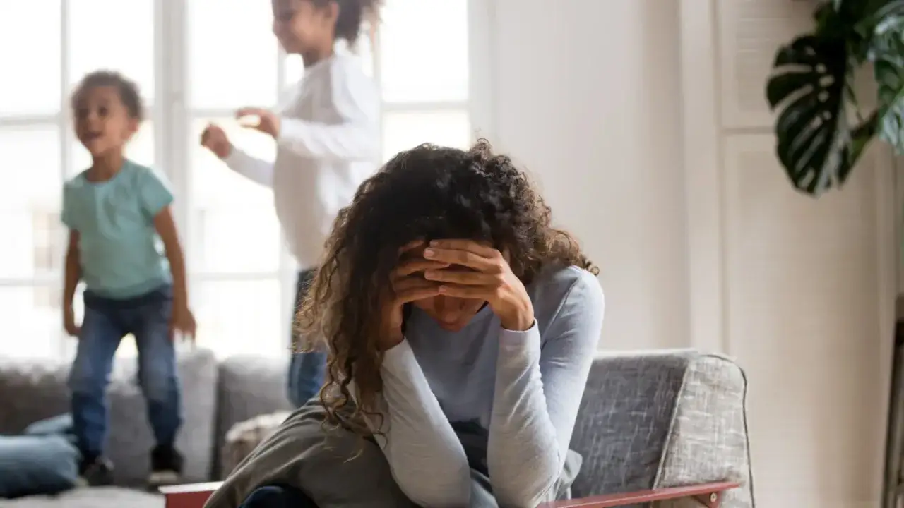 Frau mit H&auml;nden vor dem Gesicht, w&auml;hrend Kinder im Hintergrund spielen. Ein Bild, das die Herausforderungen der Pflege bei Depression verdeutlicht.