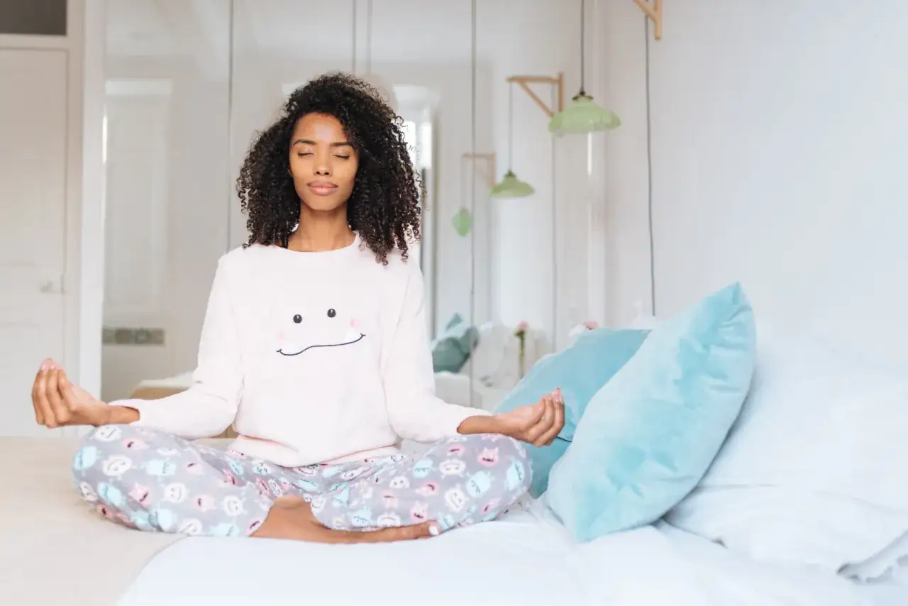 Eine Frau meditiert im Lotussitz auf einem Bett. Sie tr&auml;gt einen Pyjama mit einem l&auml;chelnden Gesicht. Sie strahlt Ruhe aus, ein Bild von zufriedenen Menschen.