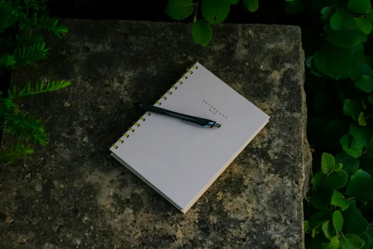 Notizbuch mit Stift auf Stein, umgeben von Gr&uuml;n. Ein Symbol f&uuml;r den Neuanfang in der Psychologie, um Gedanken und Tr&auml;ume festzuhalten.