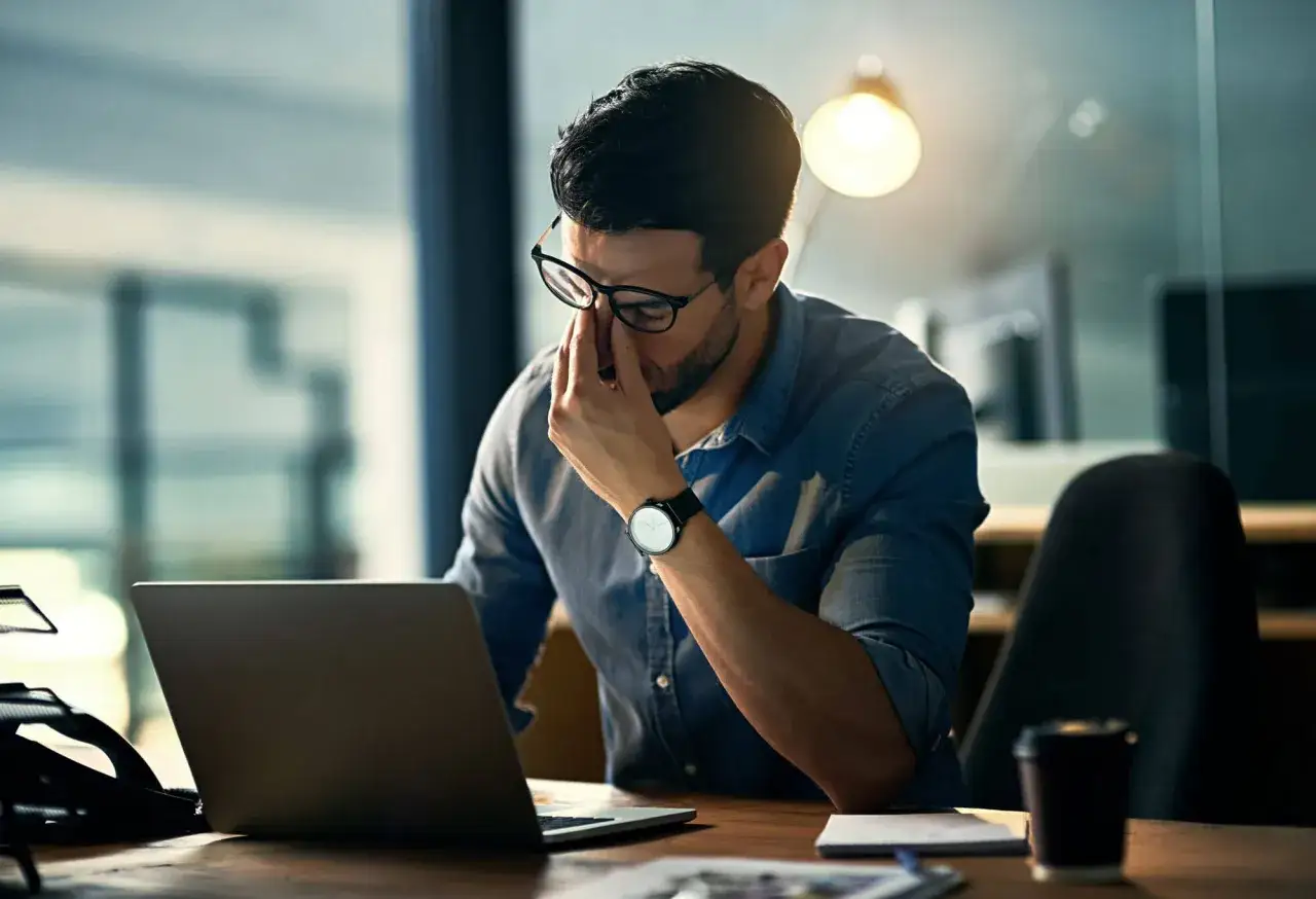 Mann mit Brille reibt sich die Augen, um den Fokus zu trainieren. Laptop und Kaffeetasse auf dem Schreibtisch.