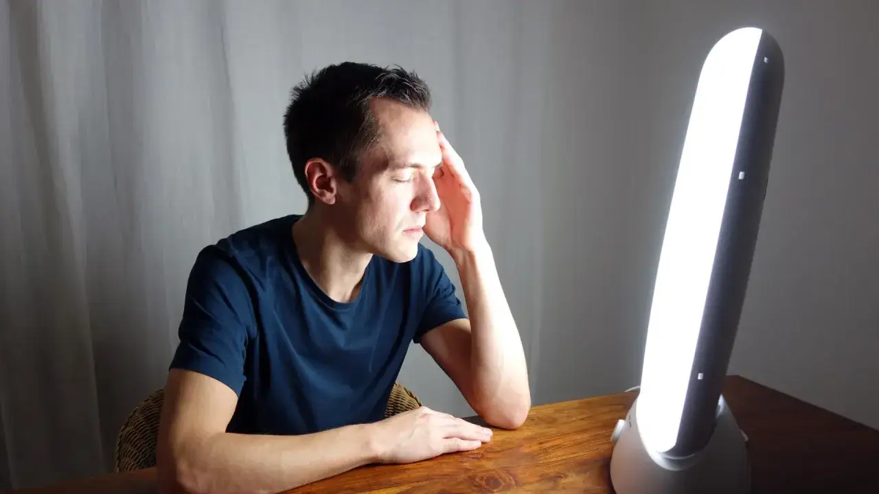 Mann mit geschlossenen Augen und Hand an der Stirn sitzt vor einer hellen Lampe, die gegen Winterdepression hilft.