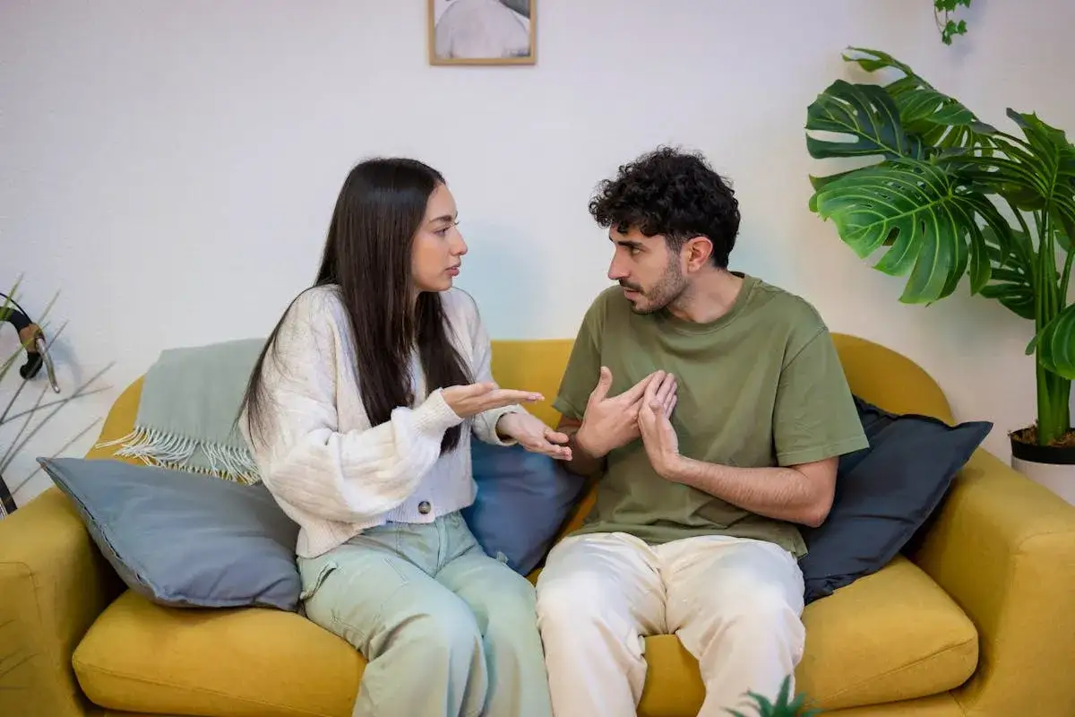 Ein Paar sitzt auf einem gelben Sofa und diskutiert angeregt. Ihre K&ouml;rpersprache deutet auf partnerschaftliche Probleme hin, w&auml;hrend sie versuchen, eine L&ouml;sung zu finden.