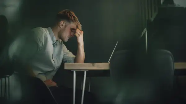 Ein Mann sitzt mit der Hand an der Stirn vor einem Laptop. Er wirkt nachdenklich, aber es ist möglich, dass er trotz Depressionen arbeitet.
