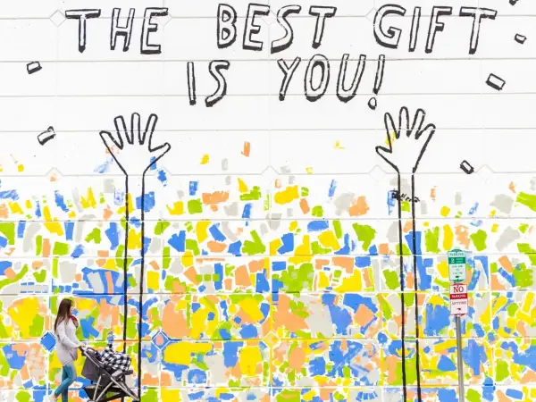 Bunte Wandmalerei mit der Botschaft "The best gift is you!". Eine Frau schiebt einen Kinderwagen. Dies erinnert an die psychologischen werte tests, die uns lehren, uns selbst wertzuschätzen.