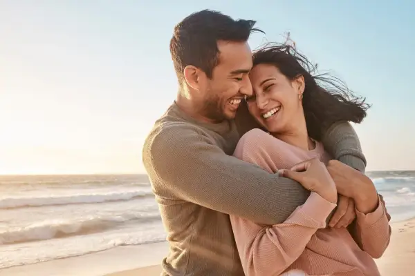 Glückliches Paar umarmt sich am Strand, lacht und genießt die Zeit. Ein perfekter Moment, um die Beziehung aufzufrischen.
