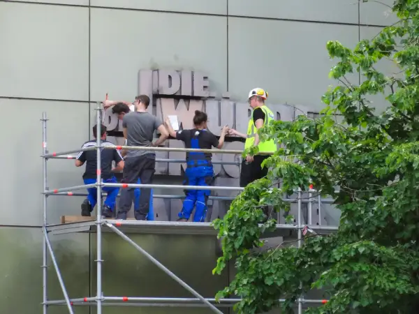 Arbeiter auf Gerüst montieren Buchstaben an einer Fassade. Ein Mensch in Warnweste gibt Anweisungen.